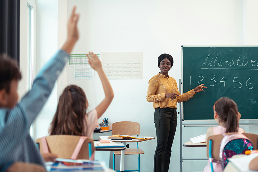 Female teacher at the front of a classroom pointing to a chalkboard as students raise their hands