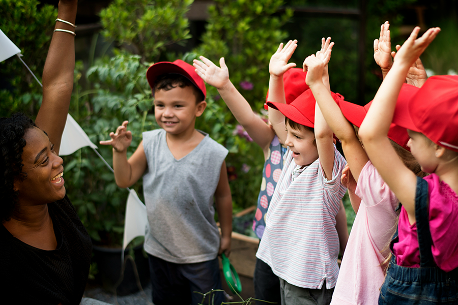 Woman and group of children wearing matching red hats smiling and raising their hands