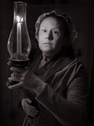 Photo of Laura F Keyes dressed as Lucy Pettengill standing in the dark holding a candle.